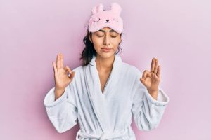 Teen girl in a white gown with sleeping mask on head above eyes, with her hands up as if meditating, and eyes closed to relax before going to sleep
